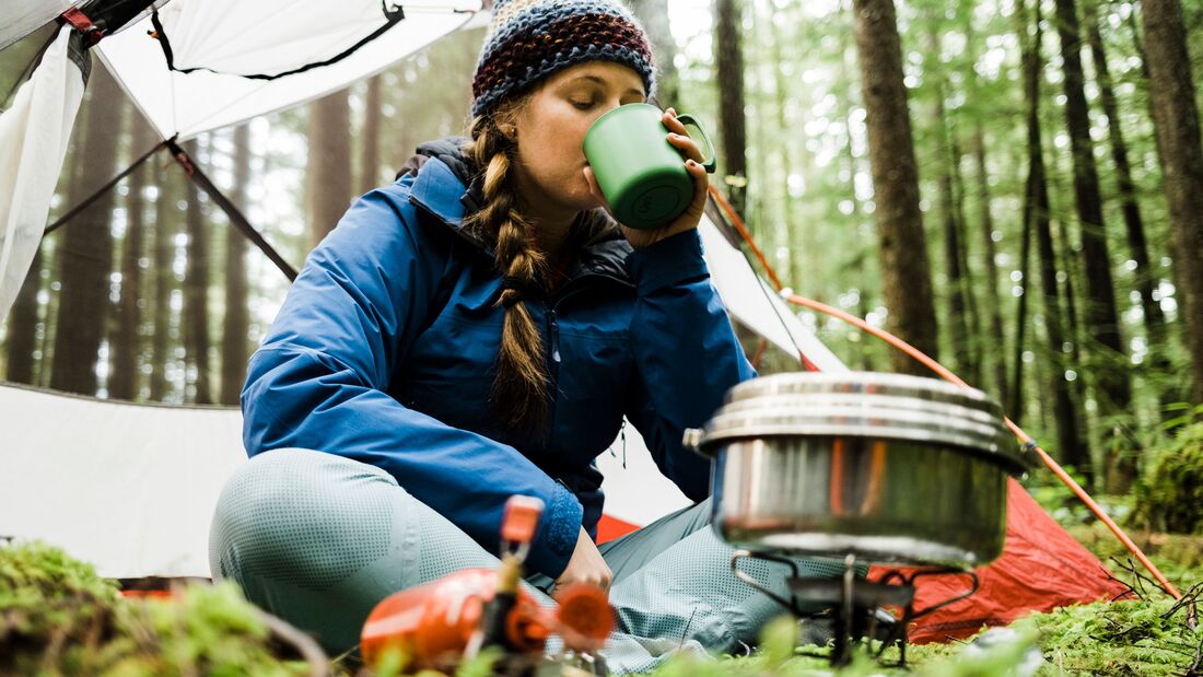 GettyImages/Alex Ratson: A blonde haired young women sits in front of her camping tent in a lush rainforest with a camp stove and pot steaming in front of her drinks a warm beverage from a mug