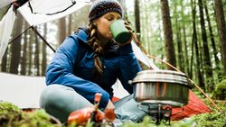 GettyImages/Alex Ratson: A blonde haired young women sits in front of her camping tent in a lush rainforest with a camp stove and pot steaming in front of her drinks a warm beverage from a mug