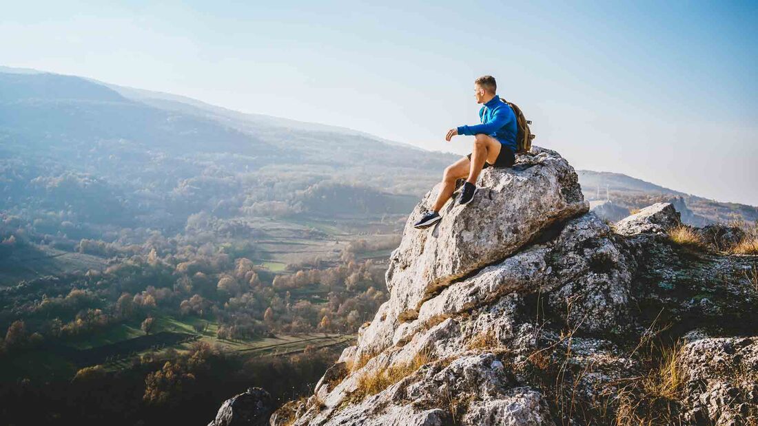 Mann der auf einem Fels sitzt und in die bergige Landschaft blickt