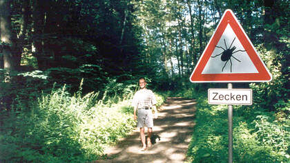 OD Spaziergänger im Wald mit Zeckenwarnschild